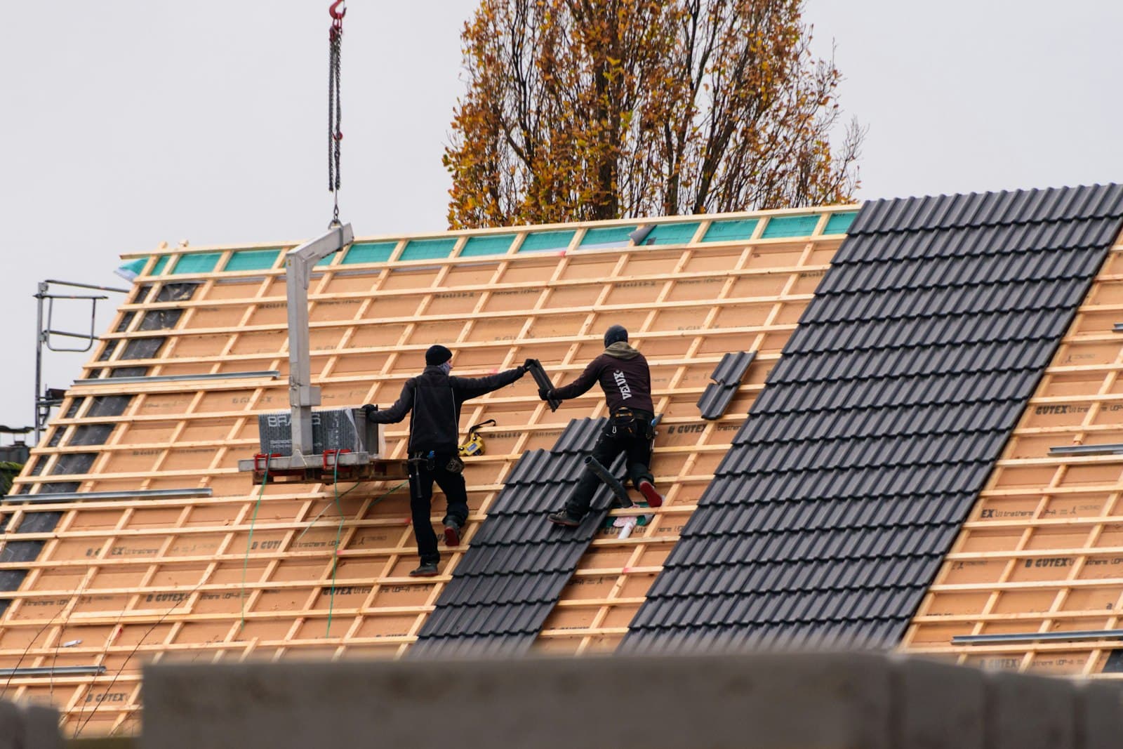 Two roofers laying crisp new clay pantile over fresh timber battens, crane in the background.